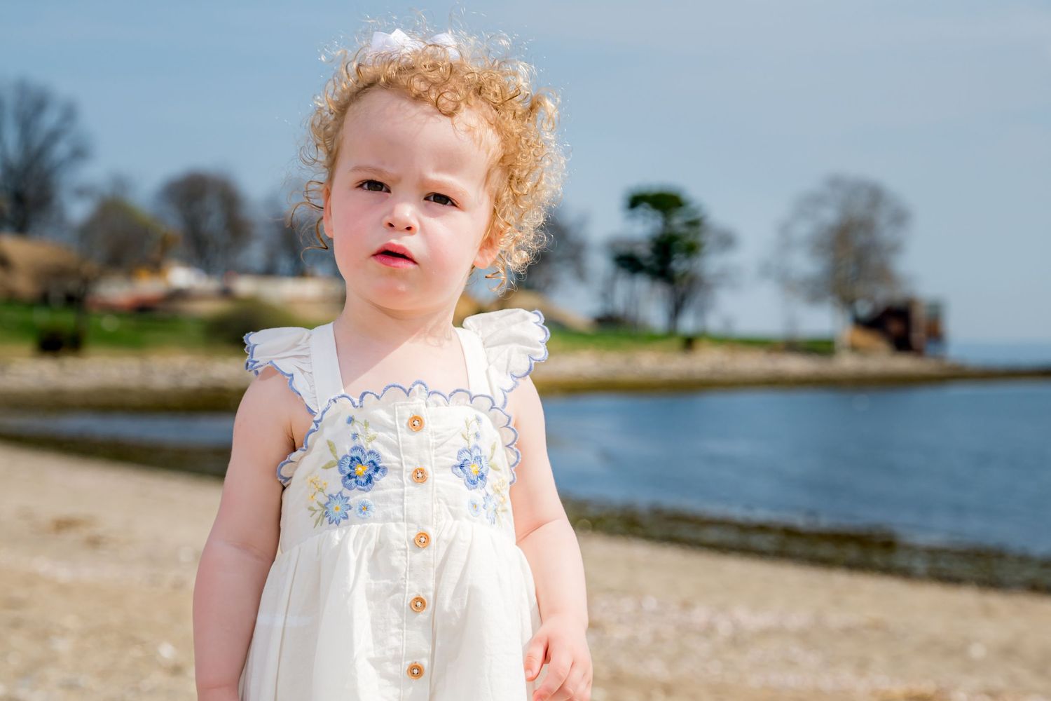 Curly-haired toddler girl in white floral sundress standing on sandy beach near calm water on sunny day.