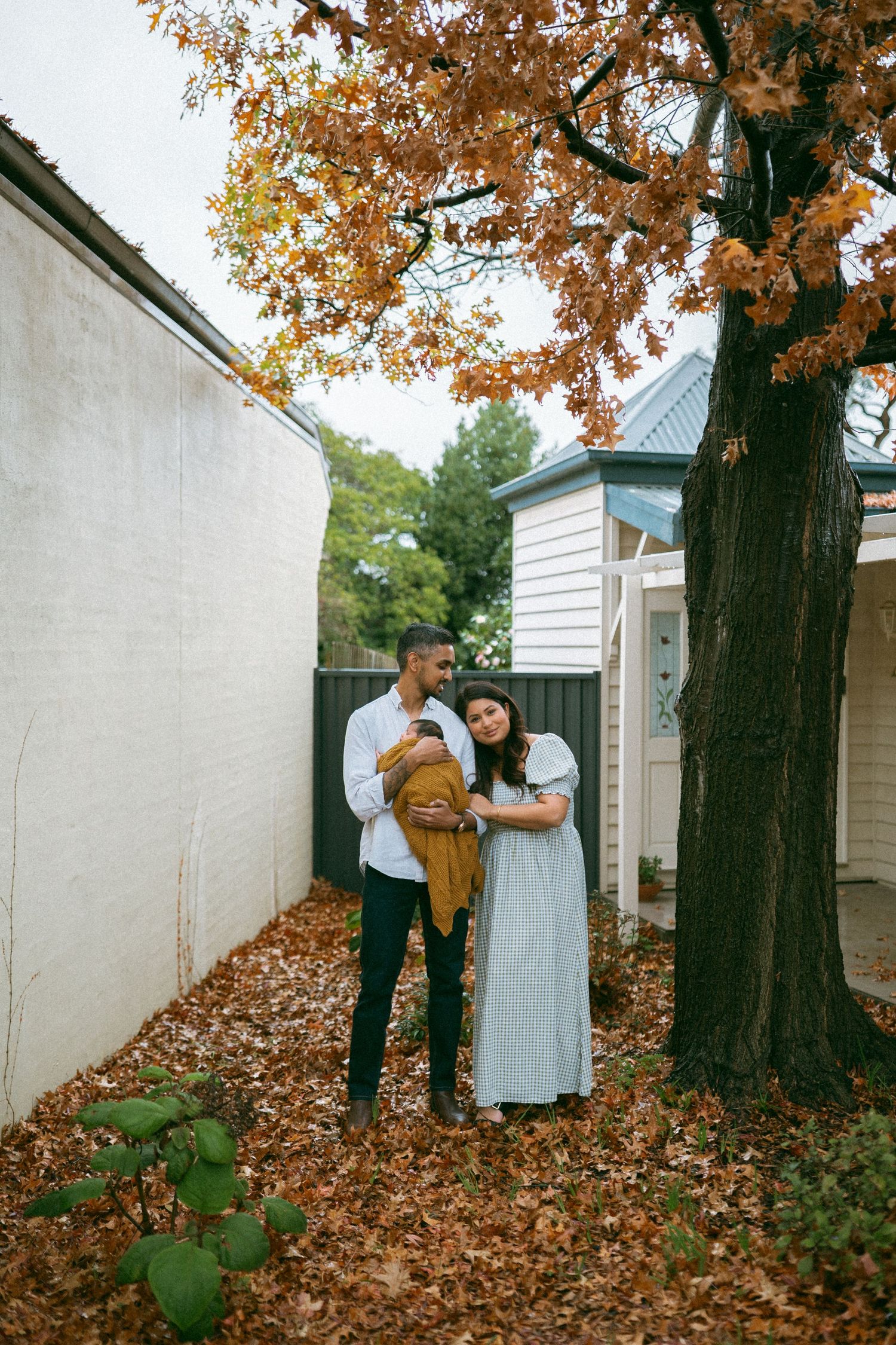 A couple stands under an autumn oak tree in a leaf-covered backyard, tenderly holding their newborn baby.