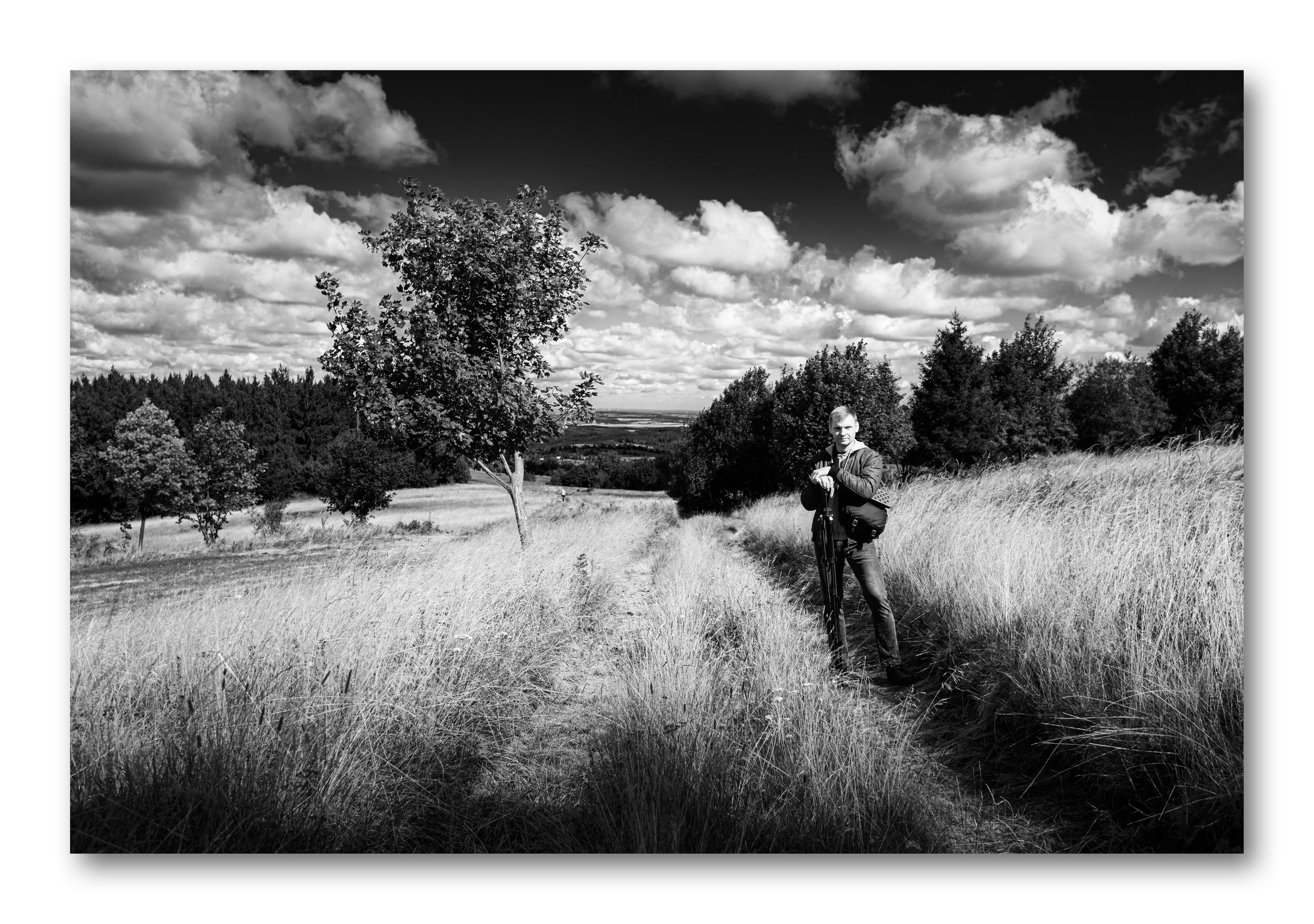 A monochrome self-portrait. A man with a backpack stands in a high, textured grass field on a slope under a dramatic cloudy sky.
