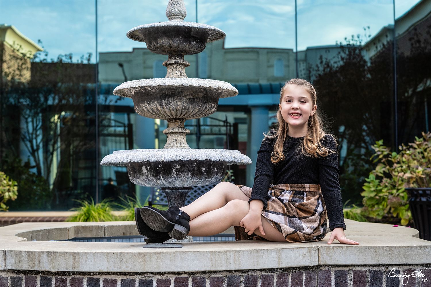 Young girl sitting on stone fountain edge outdoors, smiling in stylish outfit with plaid skirt and black top.
