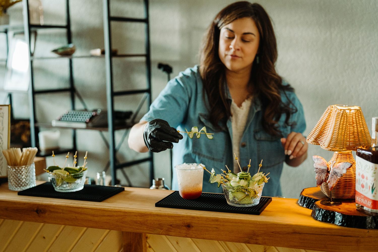 Woman in denim jacket arranging gourmet dishes on wooden bar counter with decorative lamp in background.