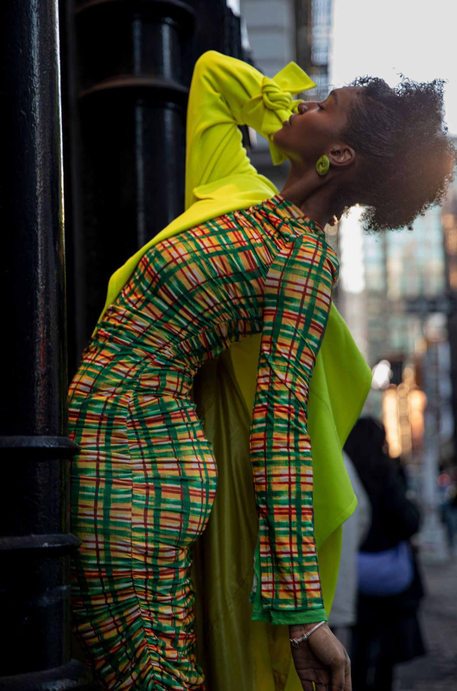 A woman in vibrant neon yellow and multicolor plaid outfit poses dramatically on a city street.