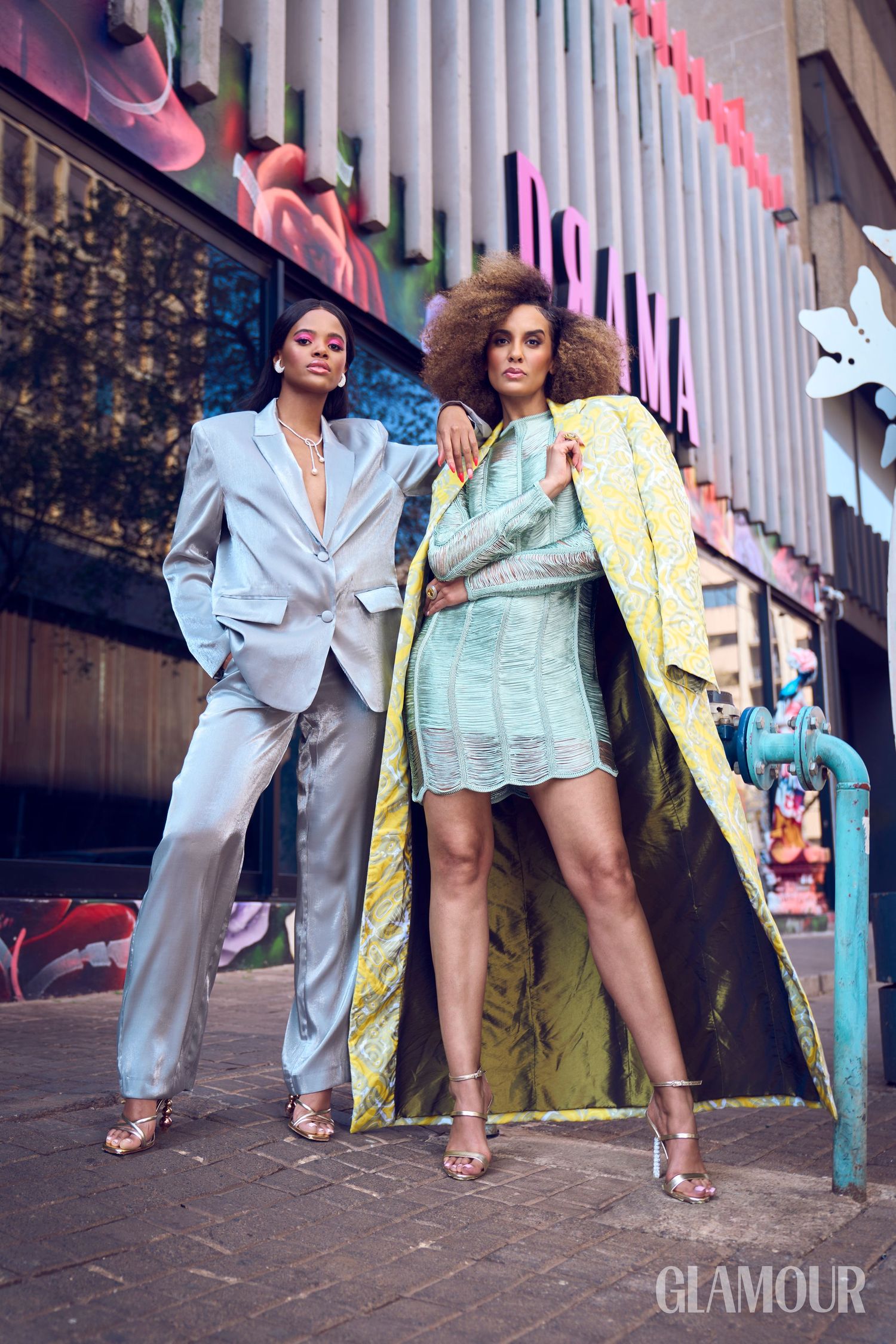 Two fashionable women in silver suit and mint dress with yellow coat pose on a city street.