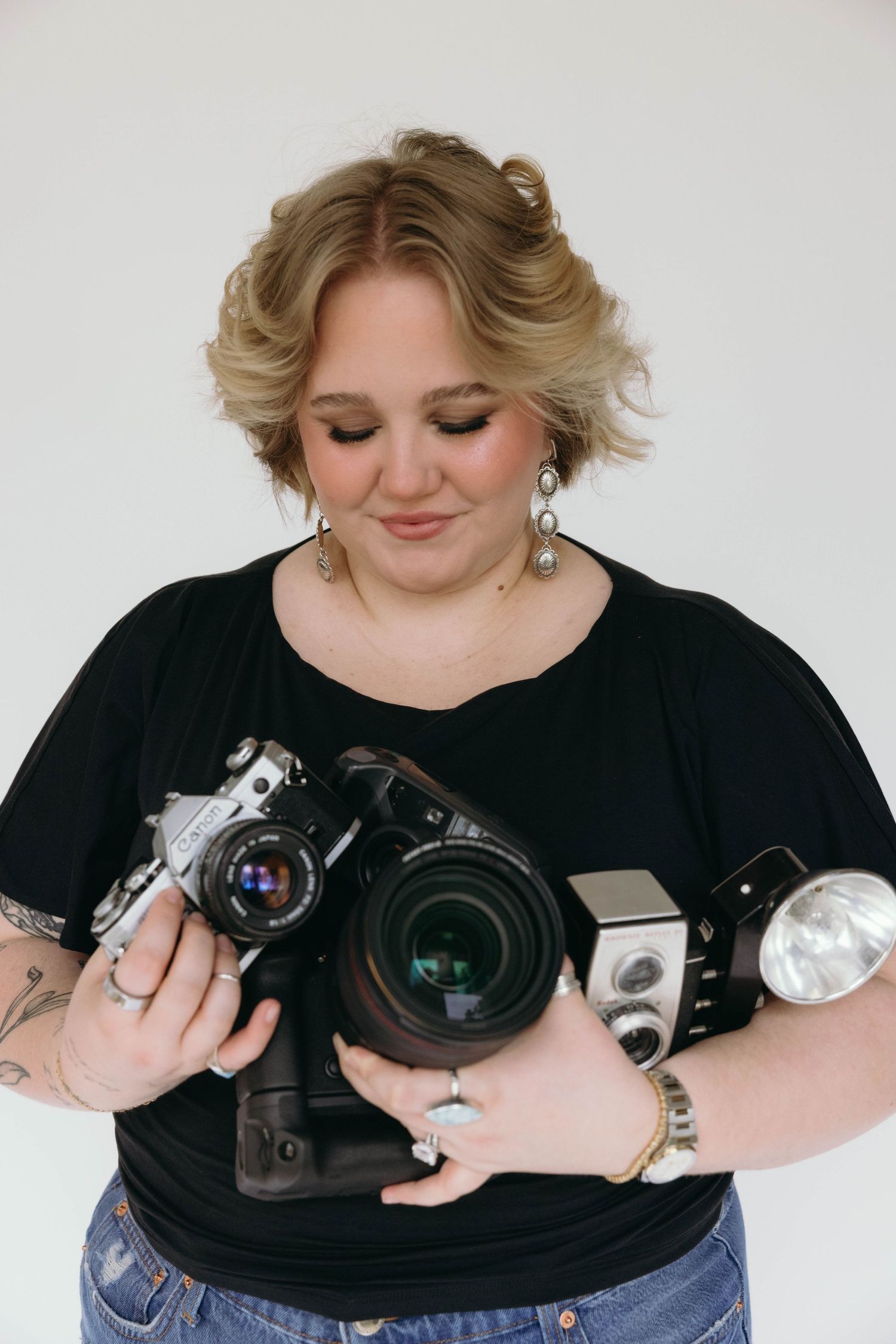 Woman in black top holding multiple cameras including film and DSLR cameras, showcasing photography equipment.