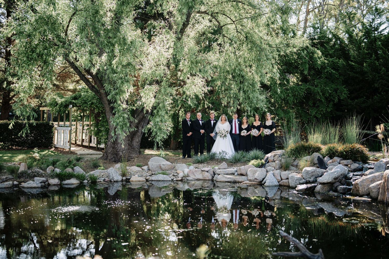 Wedding party in formal attire stands beside a rocky pond beneath a large weeping willow tree, reflected in the calm water.