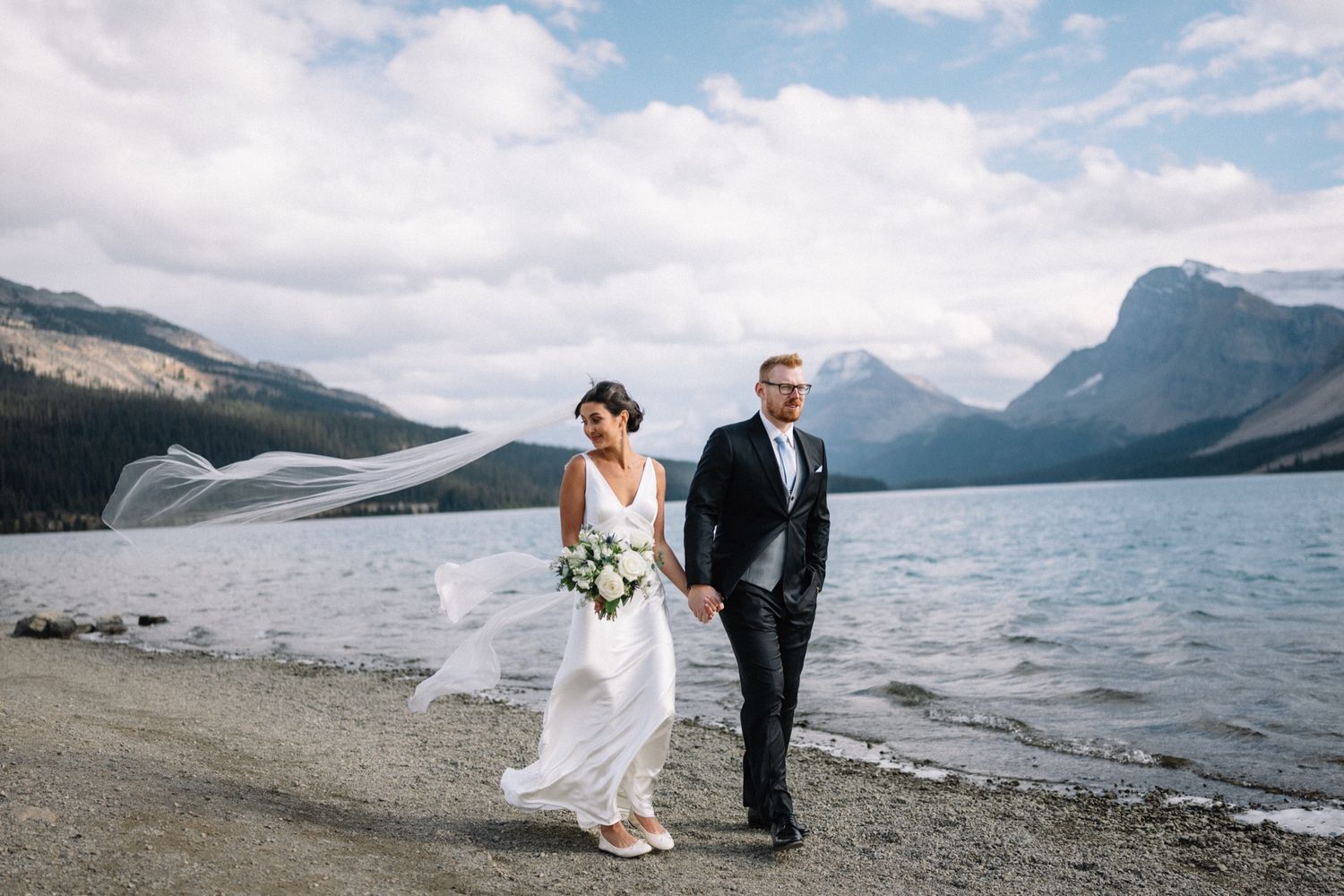 Bride and groom walking along Bow Lake near Lake Louise, veil in the wind