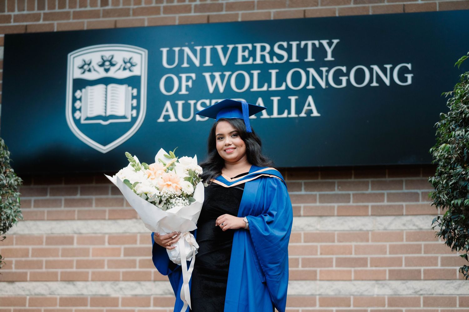 Graduate in blue gown and cap holding a white flower bouquet in front of the University of Wollongong Australia sign.