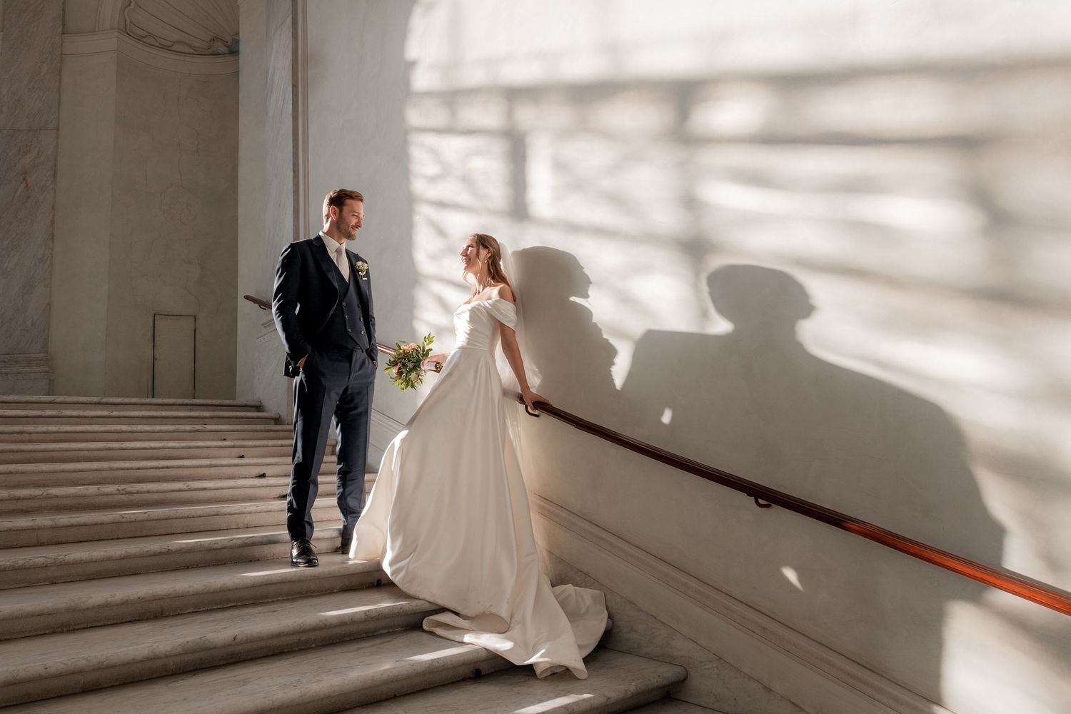 Bride in a flowing white gown and groom in a navy suit pose on grand marble stairs with dramatic window shadows cast on the wall.