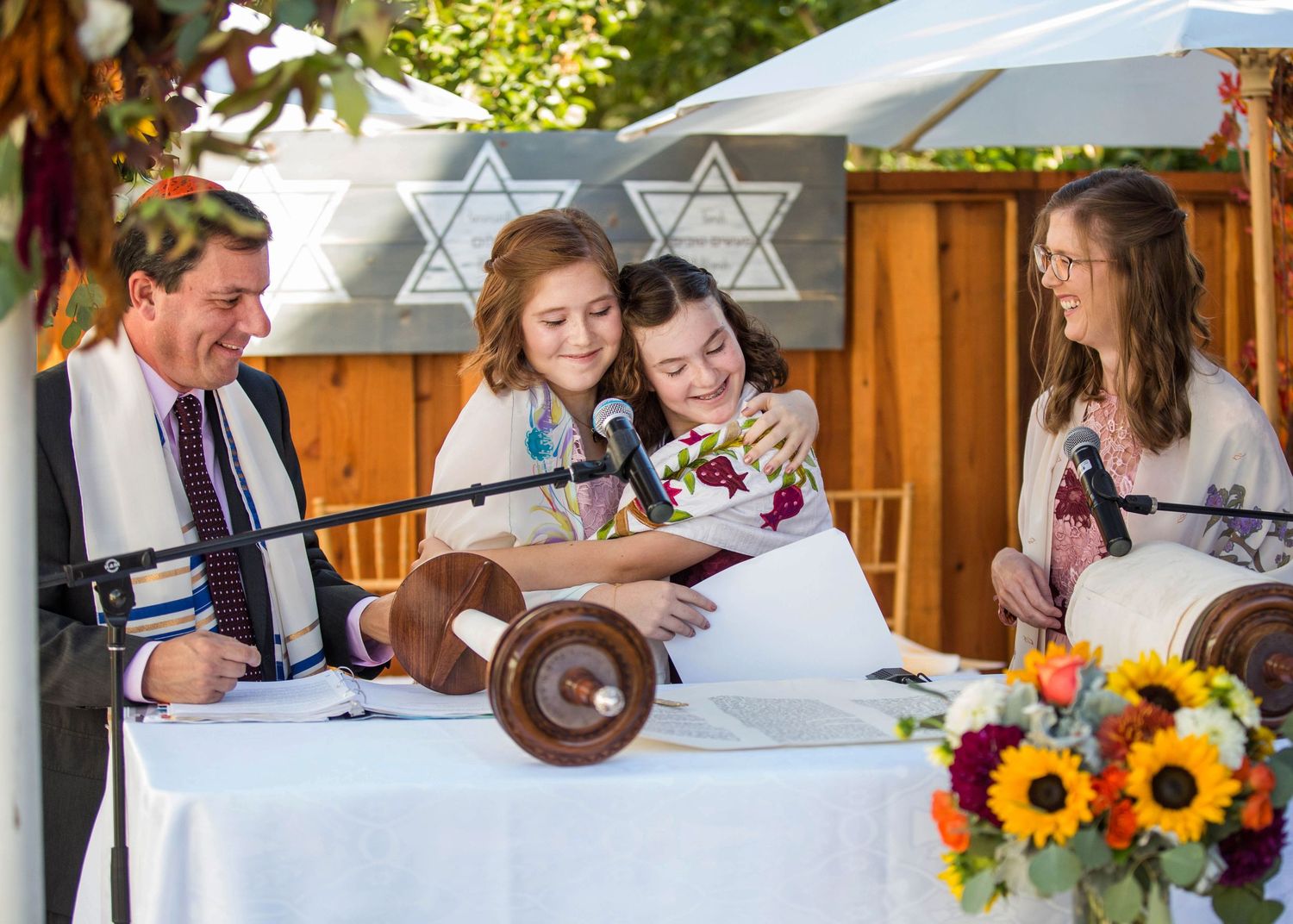 A smiling teenage boy in a tallit stands at a Torah scroll during a bar mitzvah ceremony, flanked by his mother and father in a synagogue.