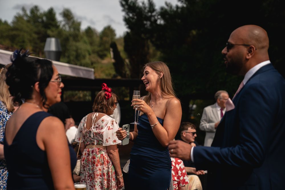 Guests in formal attire laugh and chat outdoors at a wedding reception, holding champagne glasses.