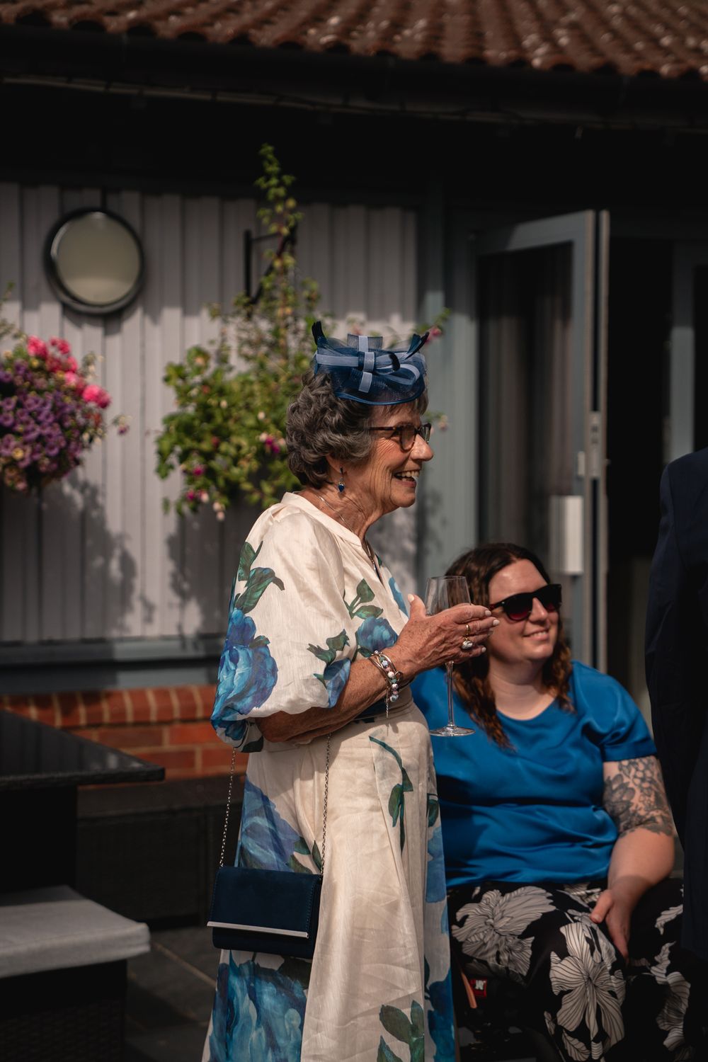 Elderly woman in floral dress and blue fascinator hat smiling outdoors at a wedding event.