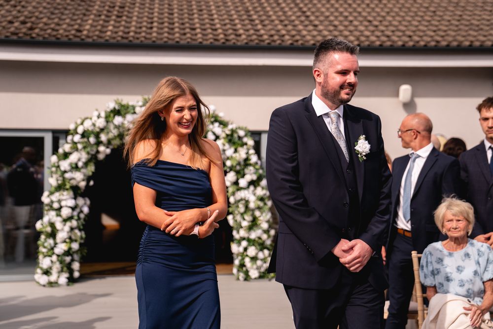 A man in a navy suit and woman in a blue dress walk together at an outdoor wedding ceremony with floral arch backdrop.