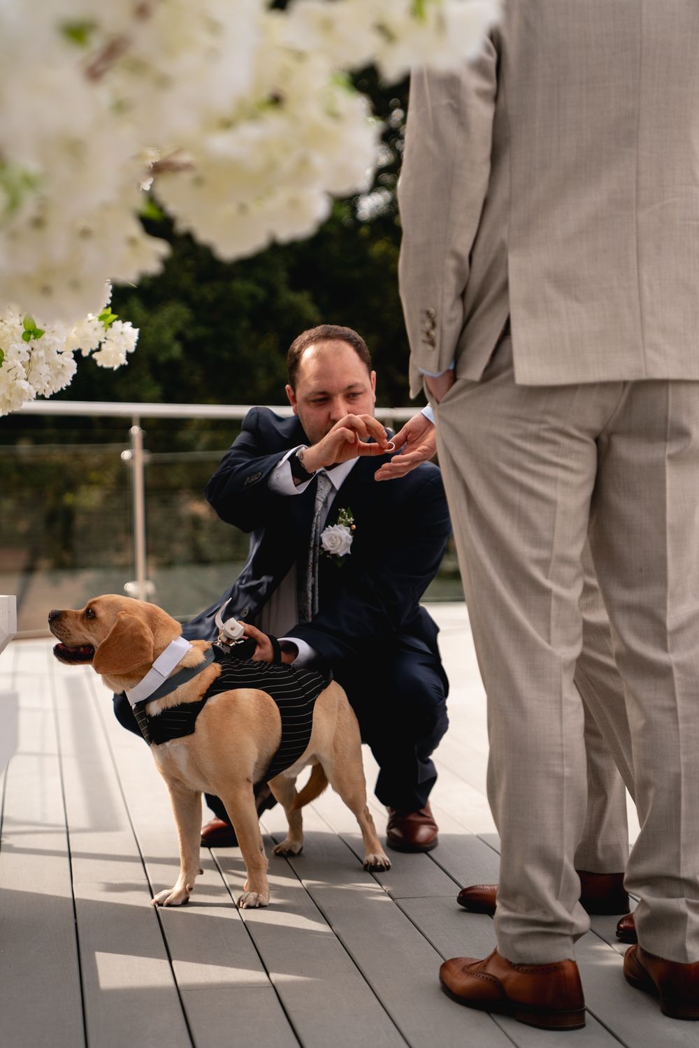 Man in navy suit crouches with dressed dog at outdoor wedding ceremony, white floral arrangements visible.