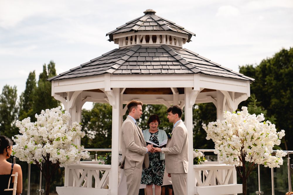 Two grooms exchange vows at a white gazebo altar adorned with white floral arrangements during an outdoor wedding ceremony.