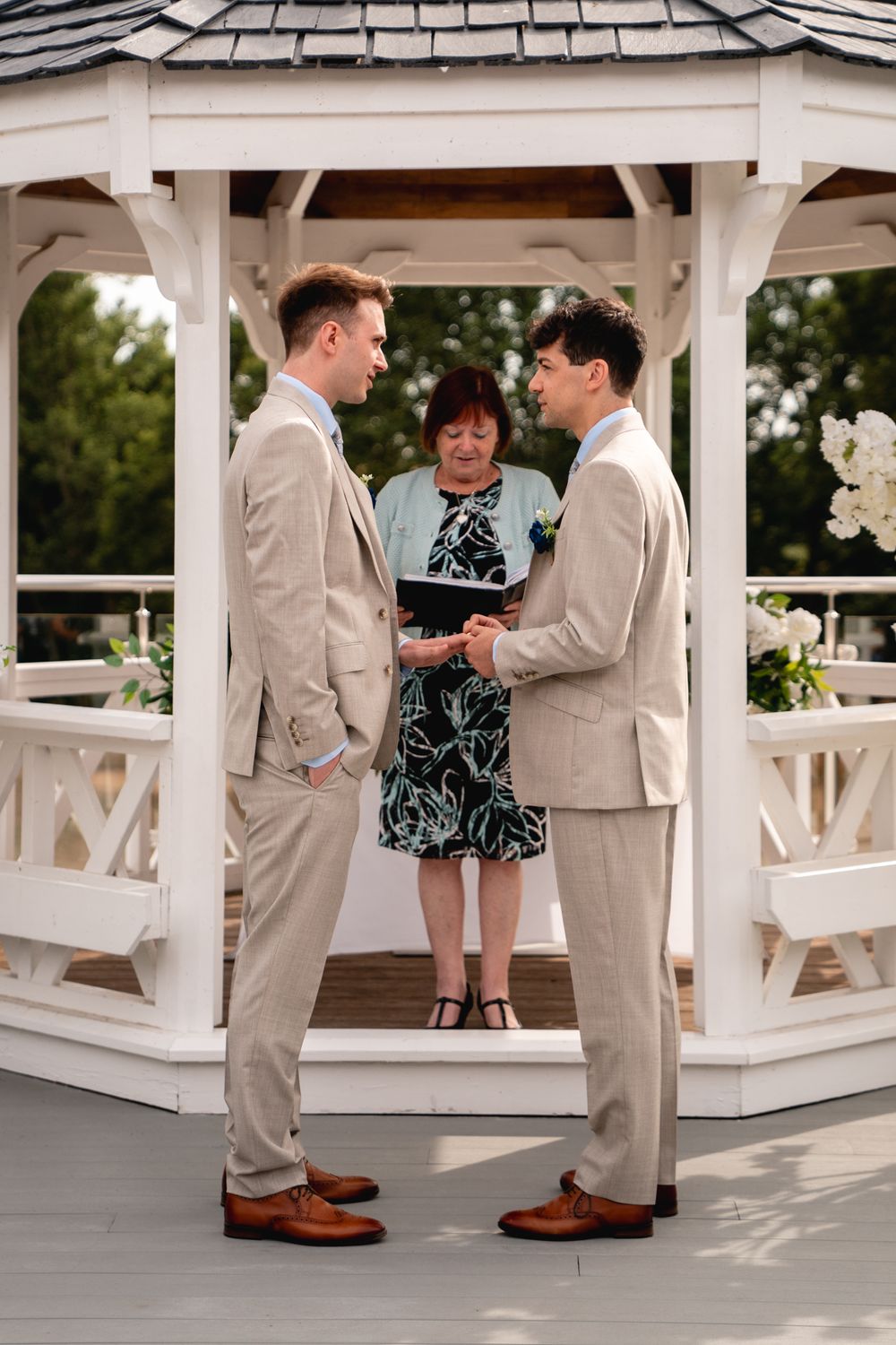 Two grooms in tan suits exchange vows under a white gazebo with a female officiant during an outdoor wedding ceremony.