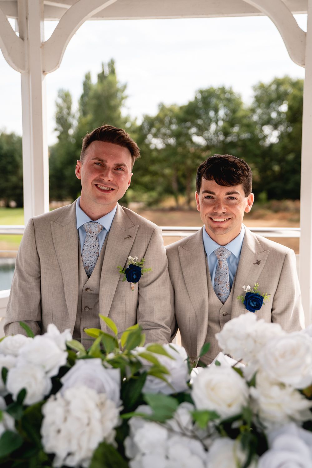 Two grooms in matching tan suits with blue boutonnieres and floral ties smile at a wedding ceremony surrounded by white flowers.