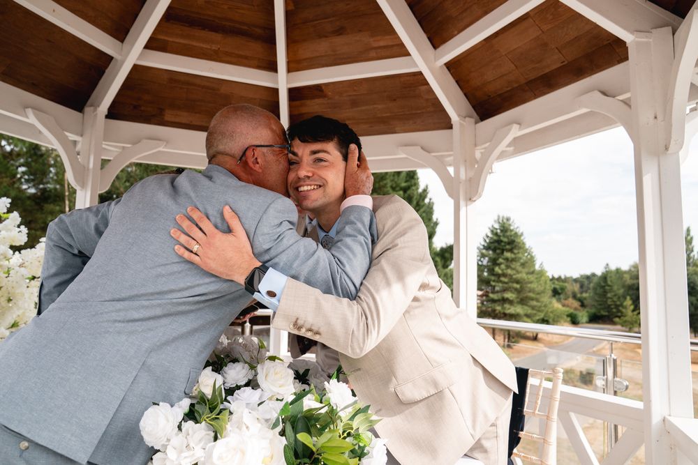 Two grooms share a heartfelt hug under a white gazebo decorated with white flowers at their wedding ceremony.