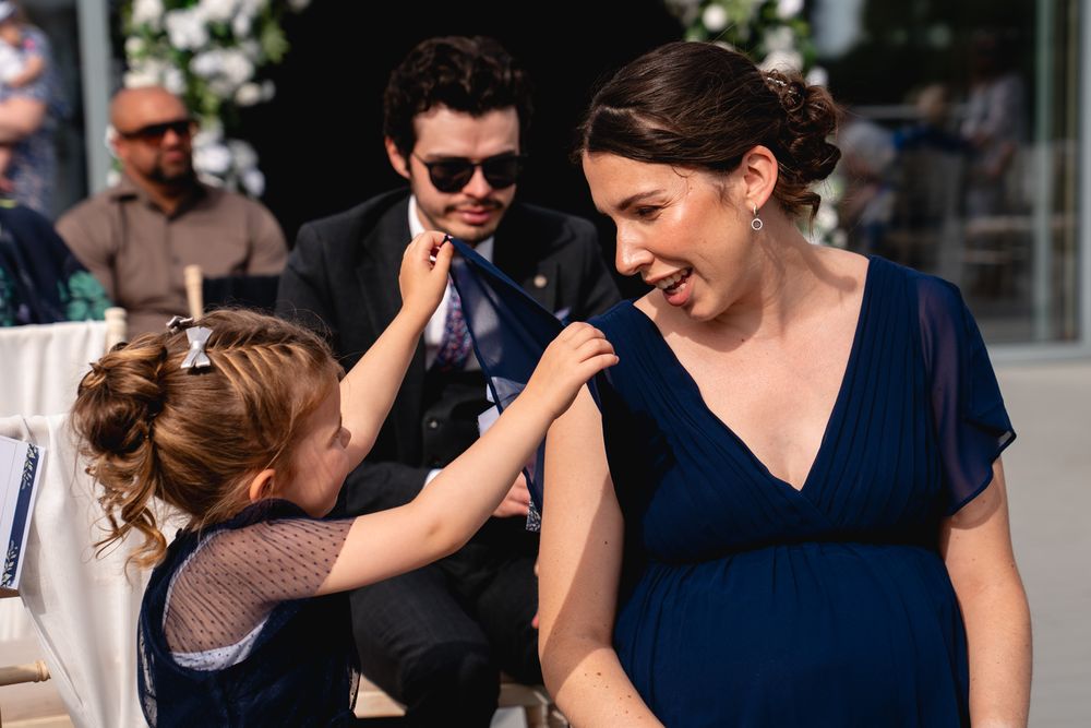 A young flower girl touches the shoulder of a smiling pregnant bridesmaid in a navy dress at an outdoor wedding.