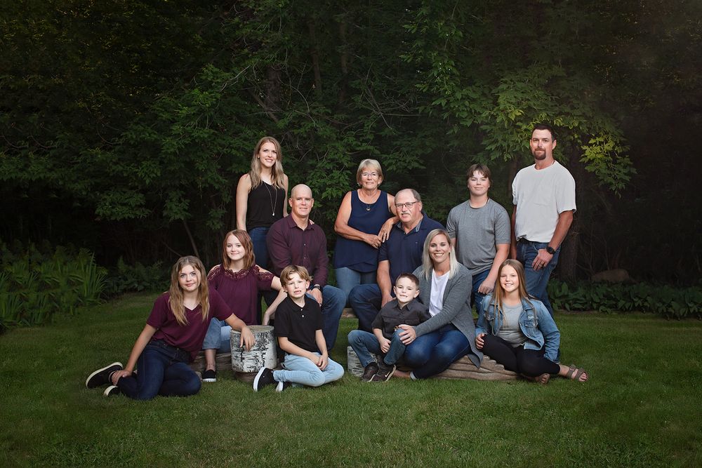 Large family of twelve, with a mix of adults, teens, and children, posed together in a grassy area with trees in behind.