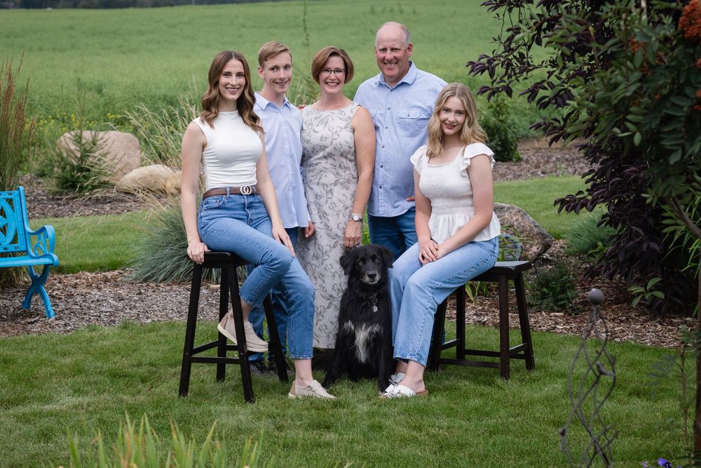 A family sits together on wooden stools in a lush green garden setting.