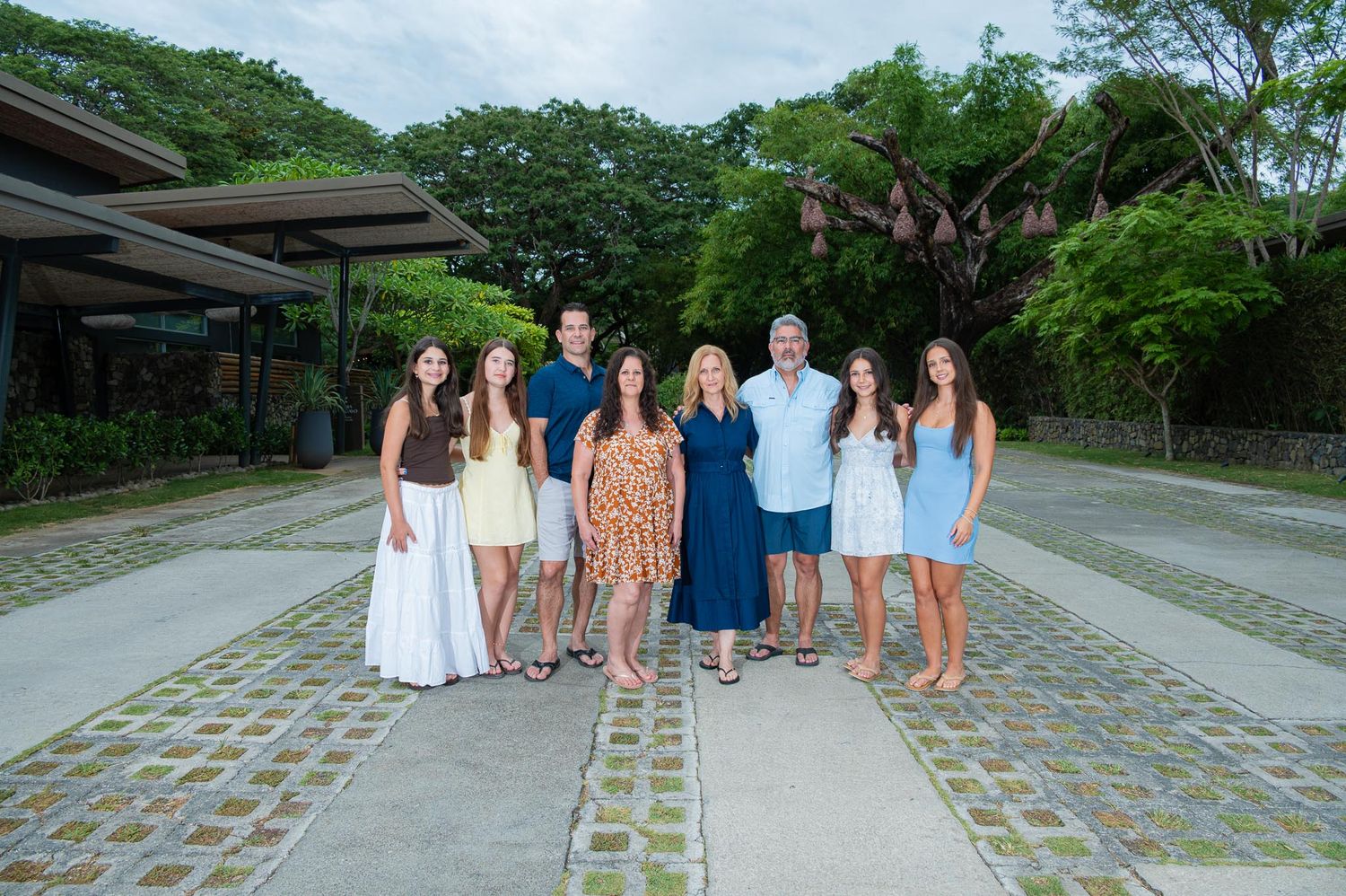 A family of eight poses together outdoors on a stone pathway surrounded by lush tropical greenery.