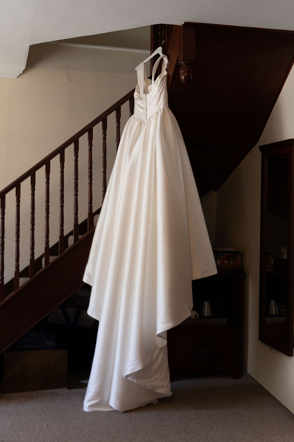 Elegant white satin wedding dress hanging on staircase banister in a home interior.
