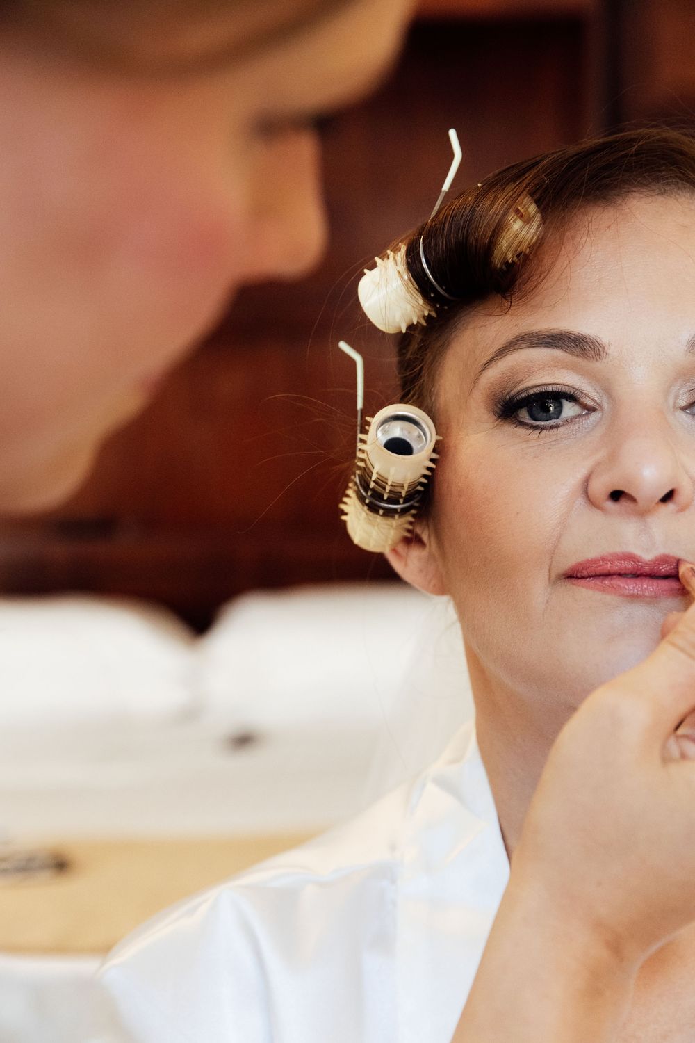 Bride getting makeup applied with hair rollers in her hair while preparing for her wedding day.