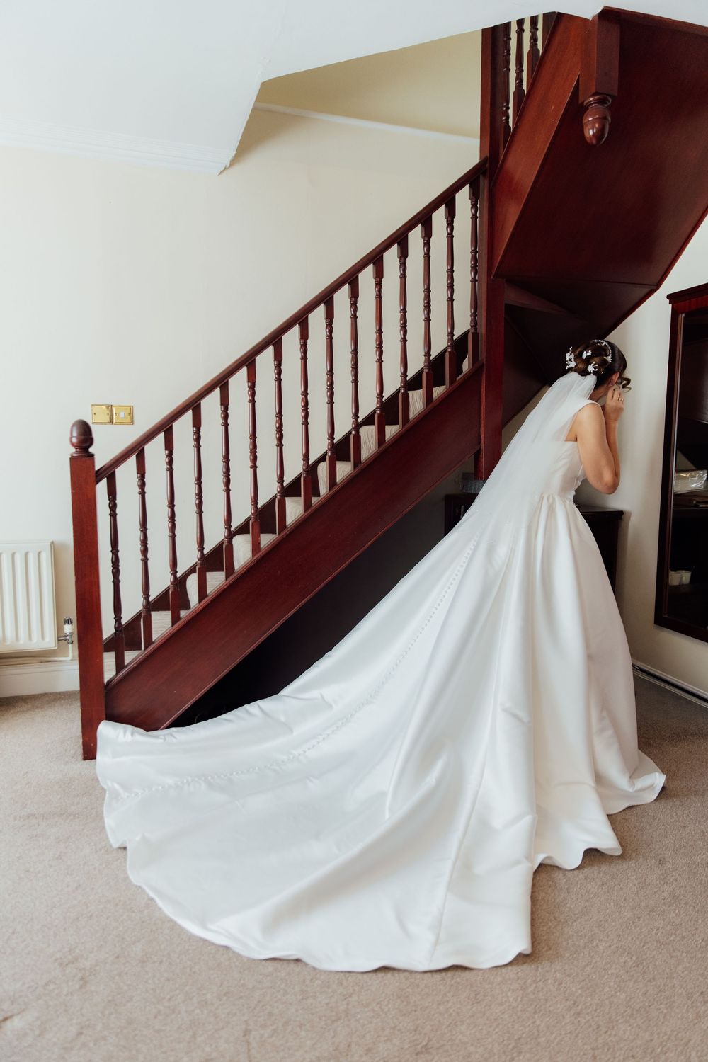 Bride in elegant white wedding gown with long train and veil standing near dark wood staircase indoors.