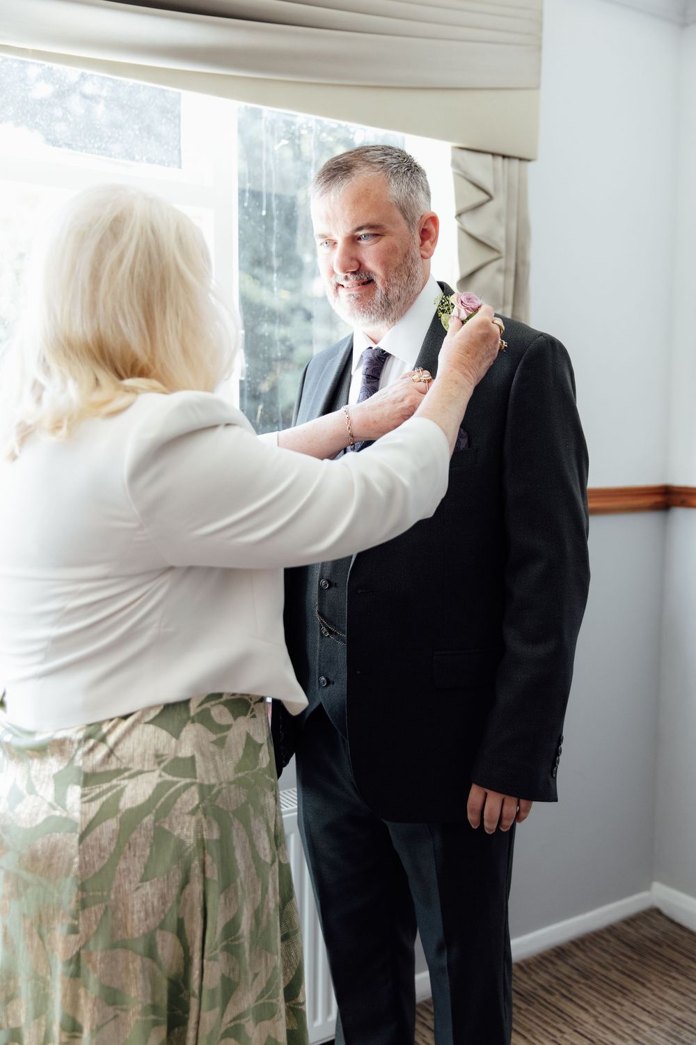 Older woman pinning a boutonniere on a smiling groom in a dark suit before a wedding ceremony.