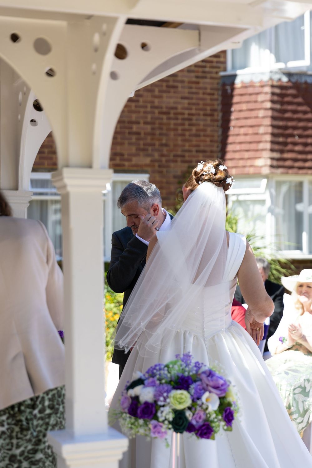 Emotional groom wiping tears as bride holds purple and white bouquet during outdoor wedding ceremony.