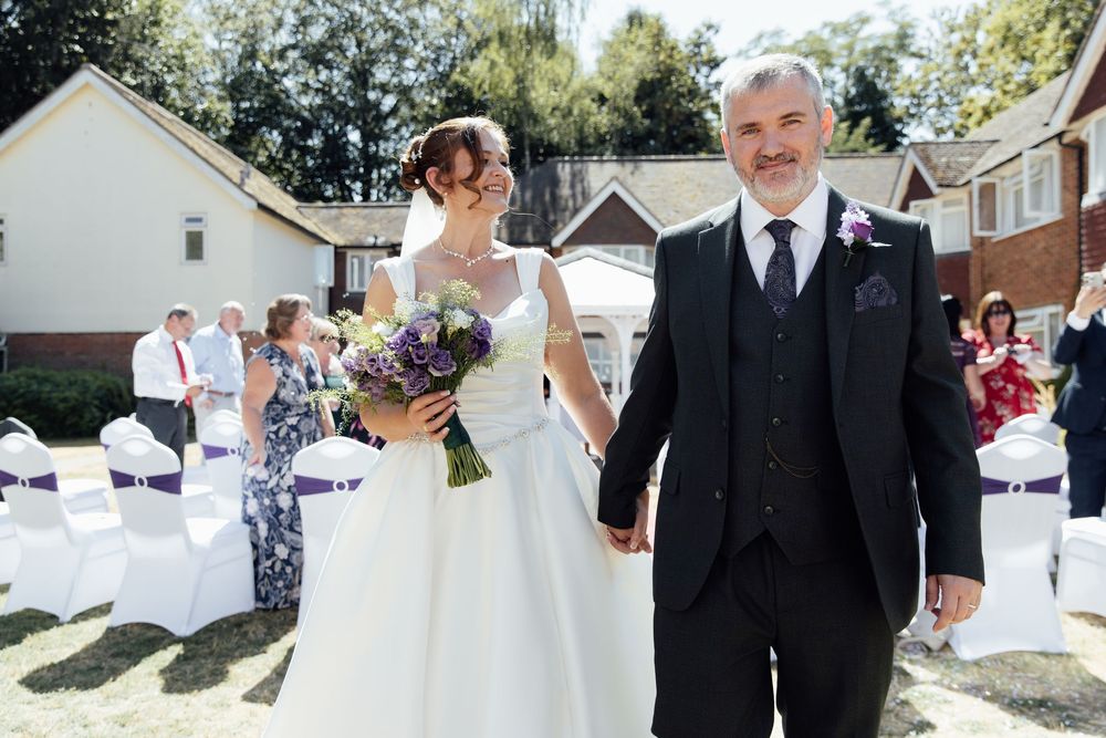 Bride and groom holding hands walking down outdoor wedding aisle, bride holding purple bouquet, sunny ceremony.