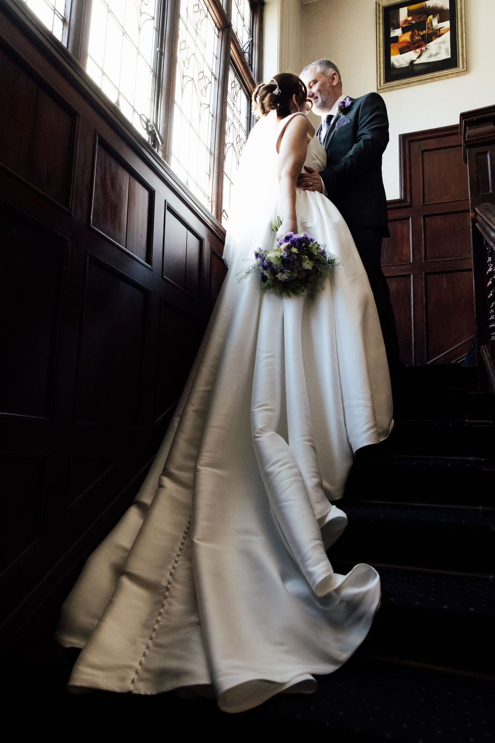 Bride and groom share a romantic moment on wooden staircase, bride holding purple bouquet in elegant white gown.
