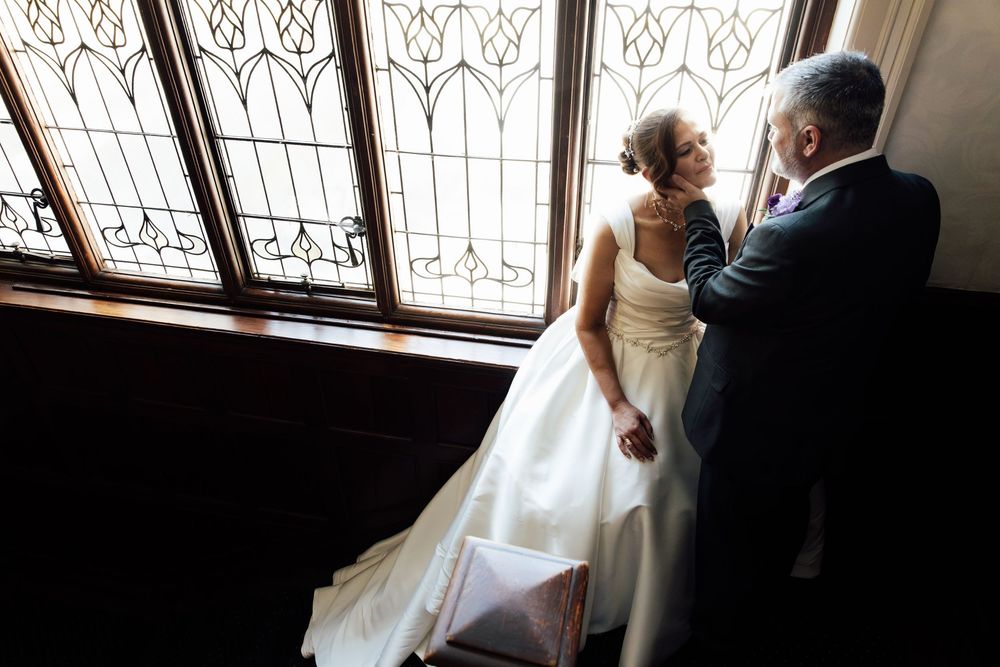 Bride and groom share a tender moment by ornate stained glass windows in elegant wedding attire.
