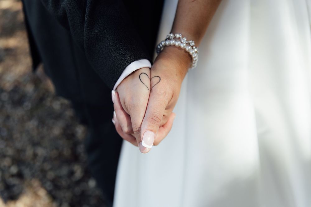 Newlyweds holding hands showing a heart tattoo, bride wearing pearl bracelet and white wedding dress.