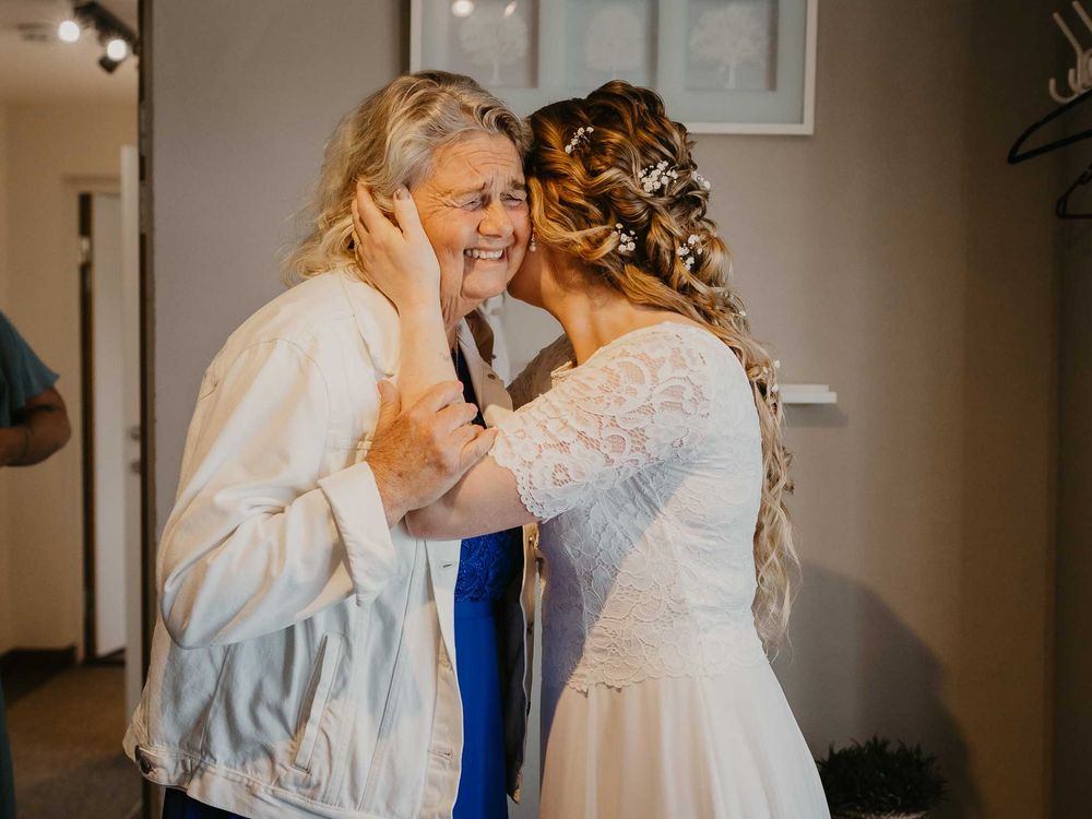Bride in white lace gown sharing a tender kiss with an older woman in a white jacket at a wedding.