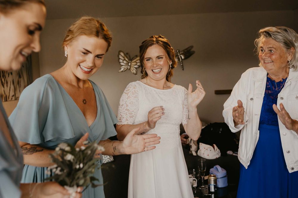 Bride in white dress laughing with bridesmaids in blue and elderly woman clapping during wedding preparations indoors.