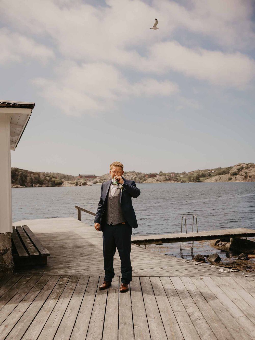 A man in a suit stands on a wooden dock by a calm lake, holding a flower boutonniere on a cloudy day.