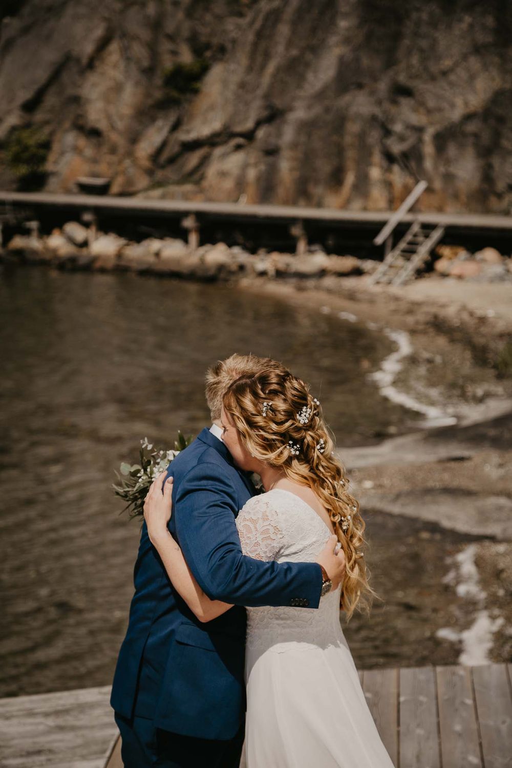 Bride and groom embracing on a wooden dock by the water, bride wearing lace dress with braided hair adorned with flowers.