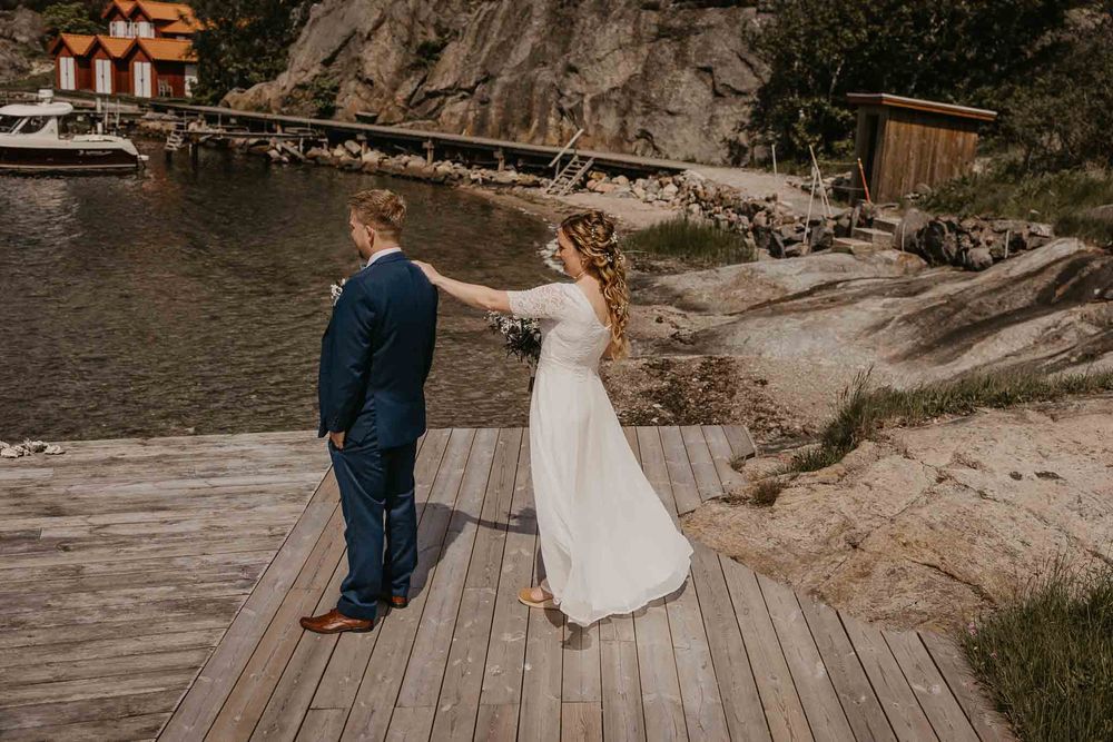 Bride and groom holding hands on a wooden dock by a rocky shoreline in a scenic outdoor wedding setting.