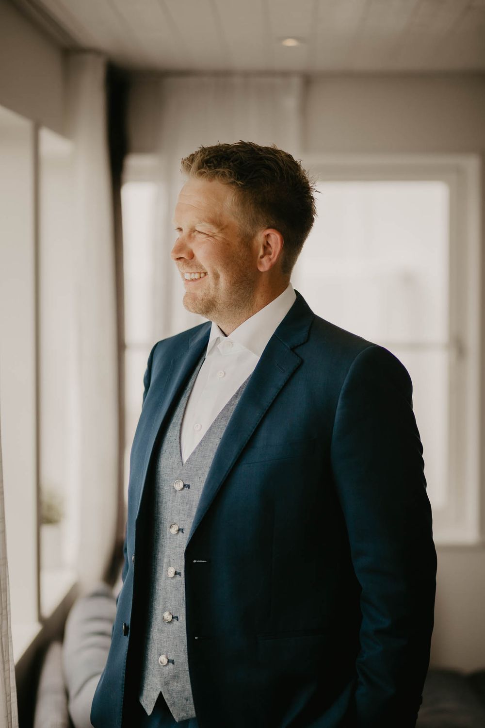 Smiling groom in navy blue suit and grey waistcoat standing by window in soft natural light.