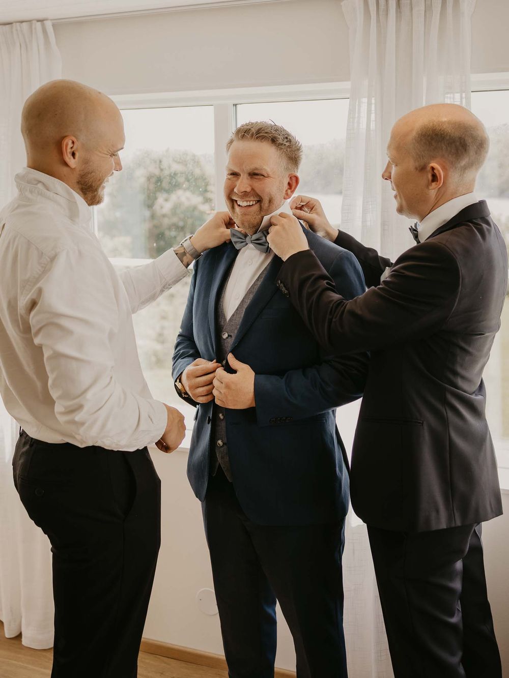 Groom in blue suit gets bow tie adjusted by two groomsmen while smiling during wedding preparations.