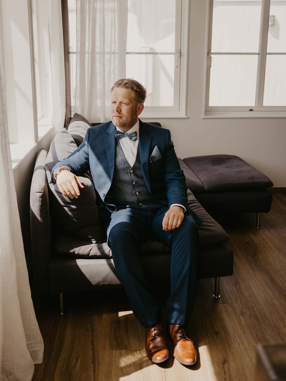 Groom in navy blue three-piece suit and brown Oxford shoes seated on sofa in natural light.