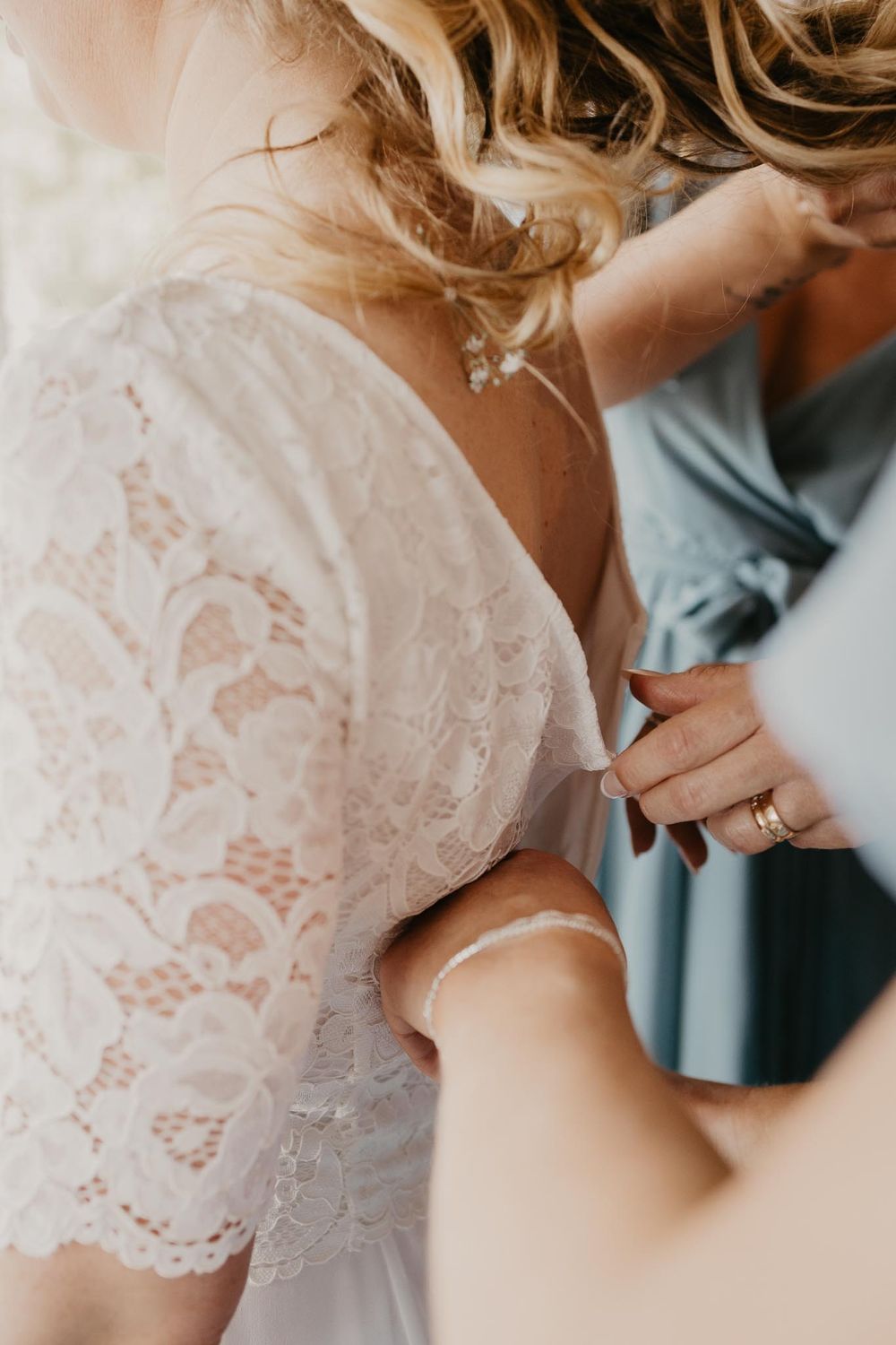 Bride in lace wedding dress being buttoned up by bridesmaid in blue dress during wedding preparations.