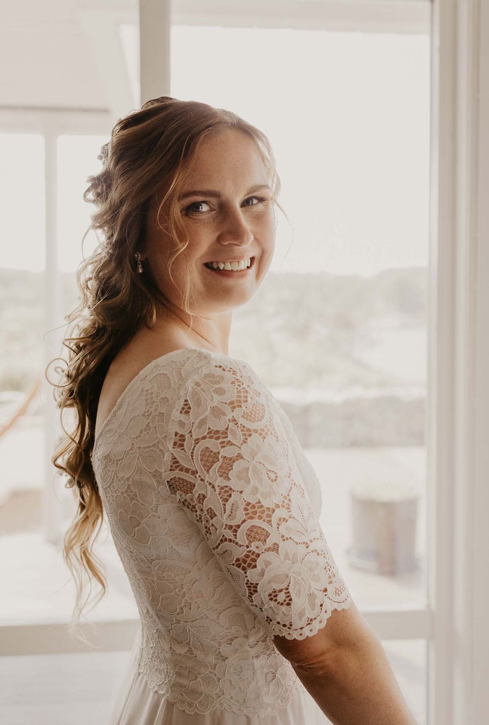 Smiling bride in white lace dress with half-up curly hairstyle standing by a bright window on her wedding day.