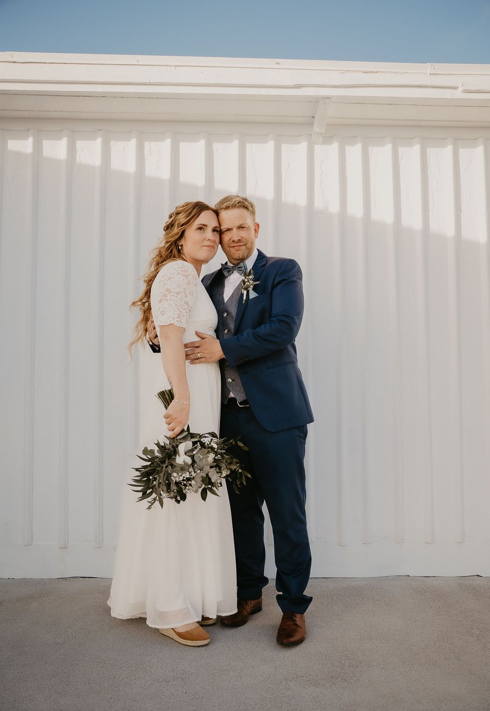 Bride in white lace dress holding greenery bouquet poses with groom in navy suit against white wall.