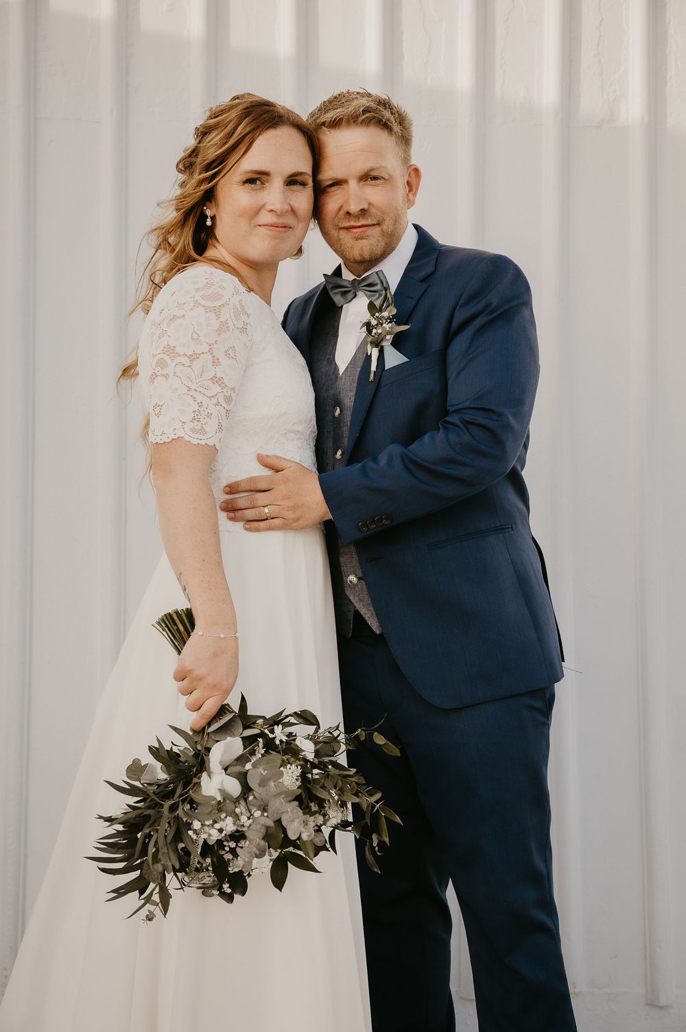 Bride in white lace dress holding greenery bouquet poses with groom in navy suit and bow tie against white backdrop.