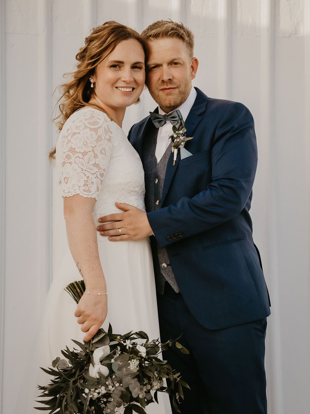 A bride and groom pose together in wedding attire, she holds a bouquet, he wears a navy suit with bow tie.