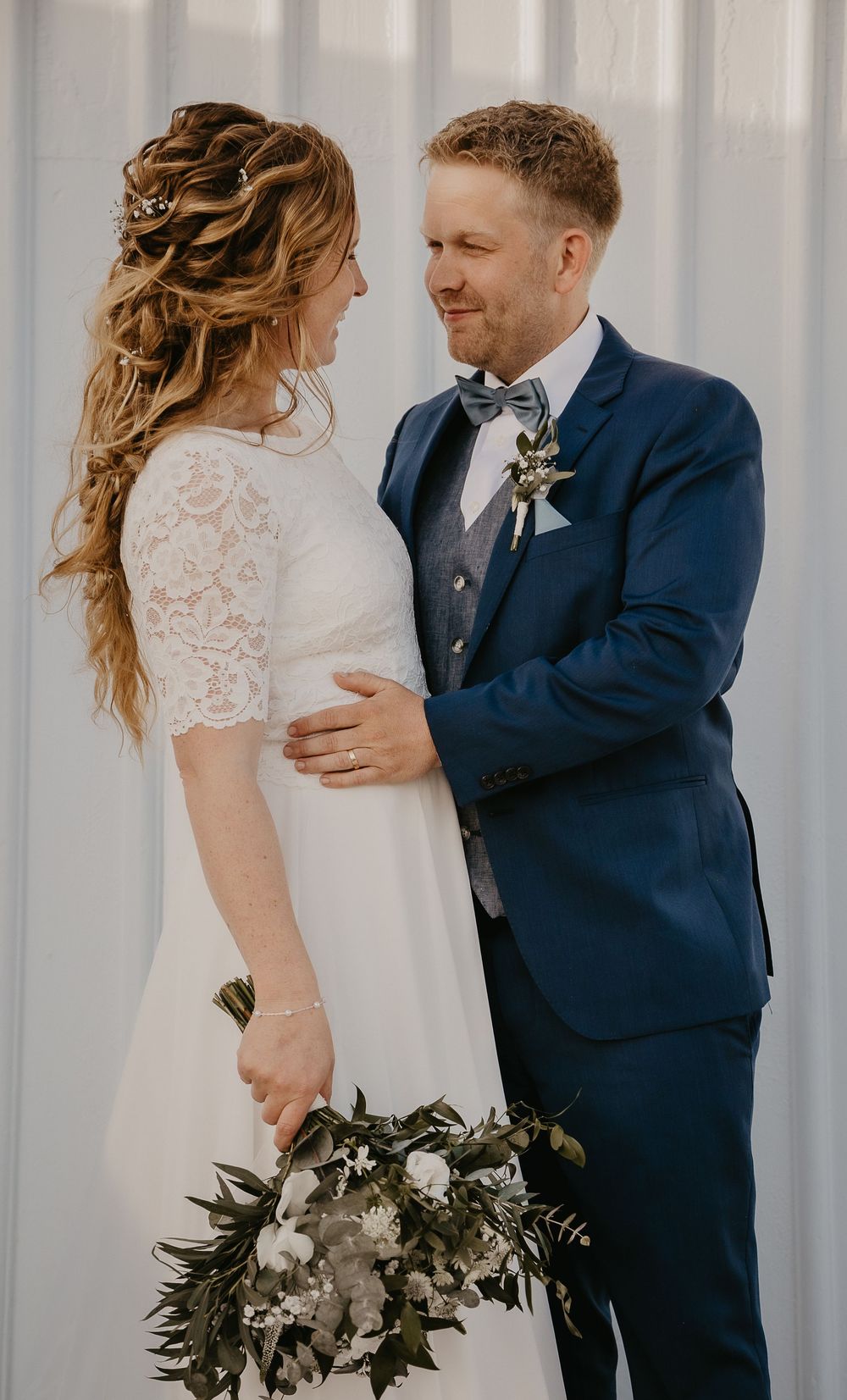 A bride in a lace dress holding a greenery bouquet gazes at her groom in a navy suit with a bow tie.
