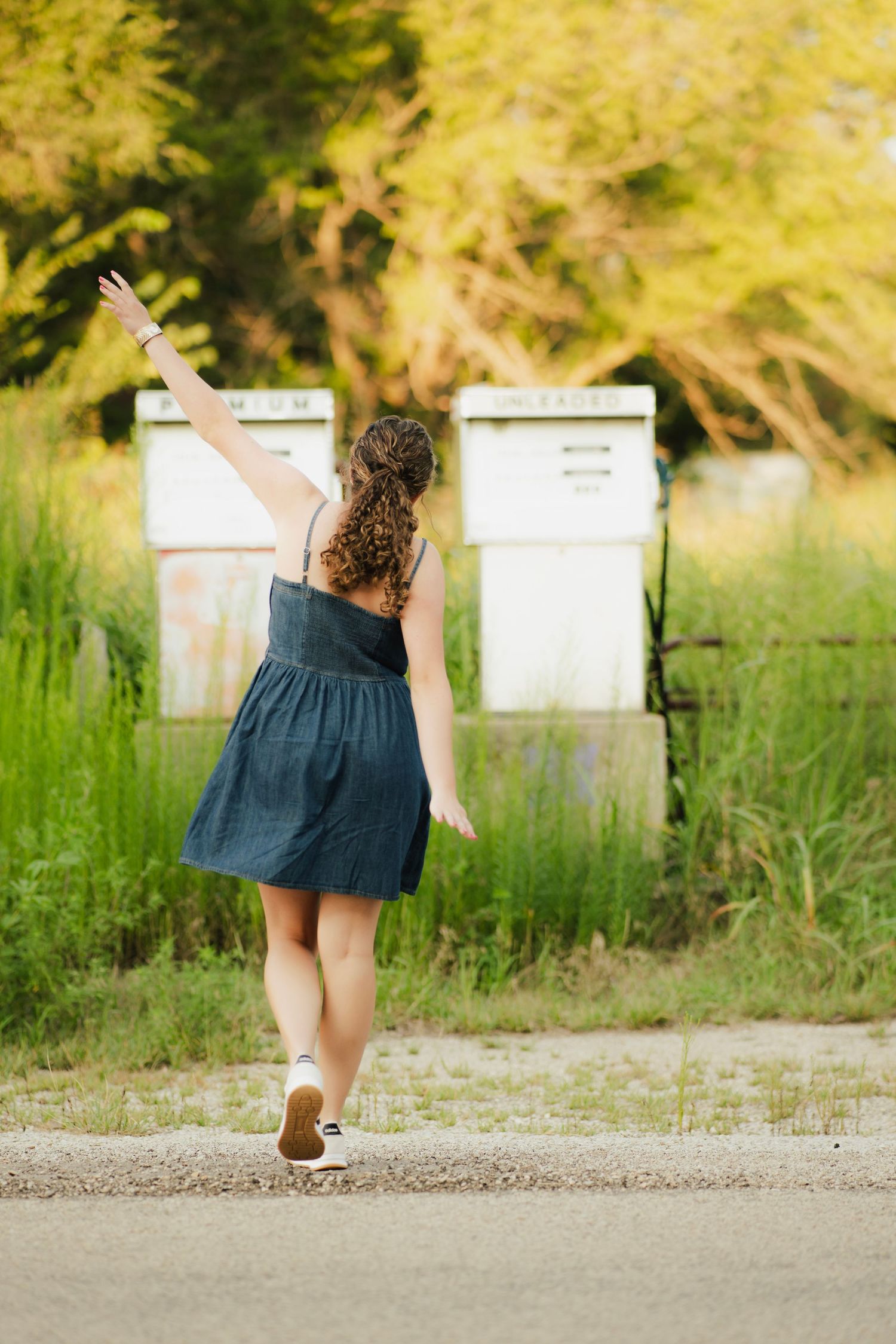 Young woman in blue dress walking toward mailboxes with arm raised, surrounded by lush green trees.