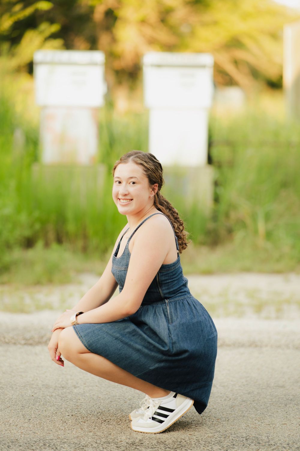 Smiling teenage girl crouching on pavement in denim dress and Adidas sneakers outdoors.